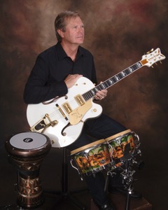 Studio portrait of Michael McCormick seated with a white Gretsch guitar beside hand drums